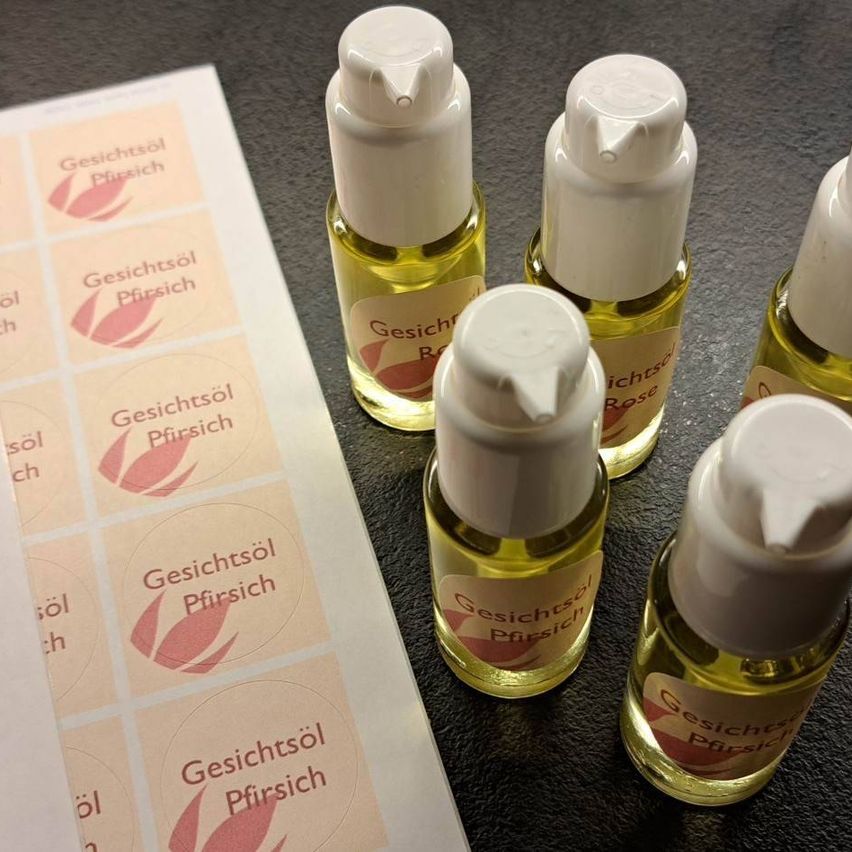 Fünf kleine Flaschen Gesichtsöl mit weißen Deckeln sind auf einem Tisch ausgestellt, begleitet von einem Etikettenbogen. Jede Flasche hat ein einzigartiges Etikett, wie 'Rose' und 'Pfirsich'.