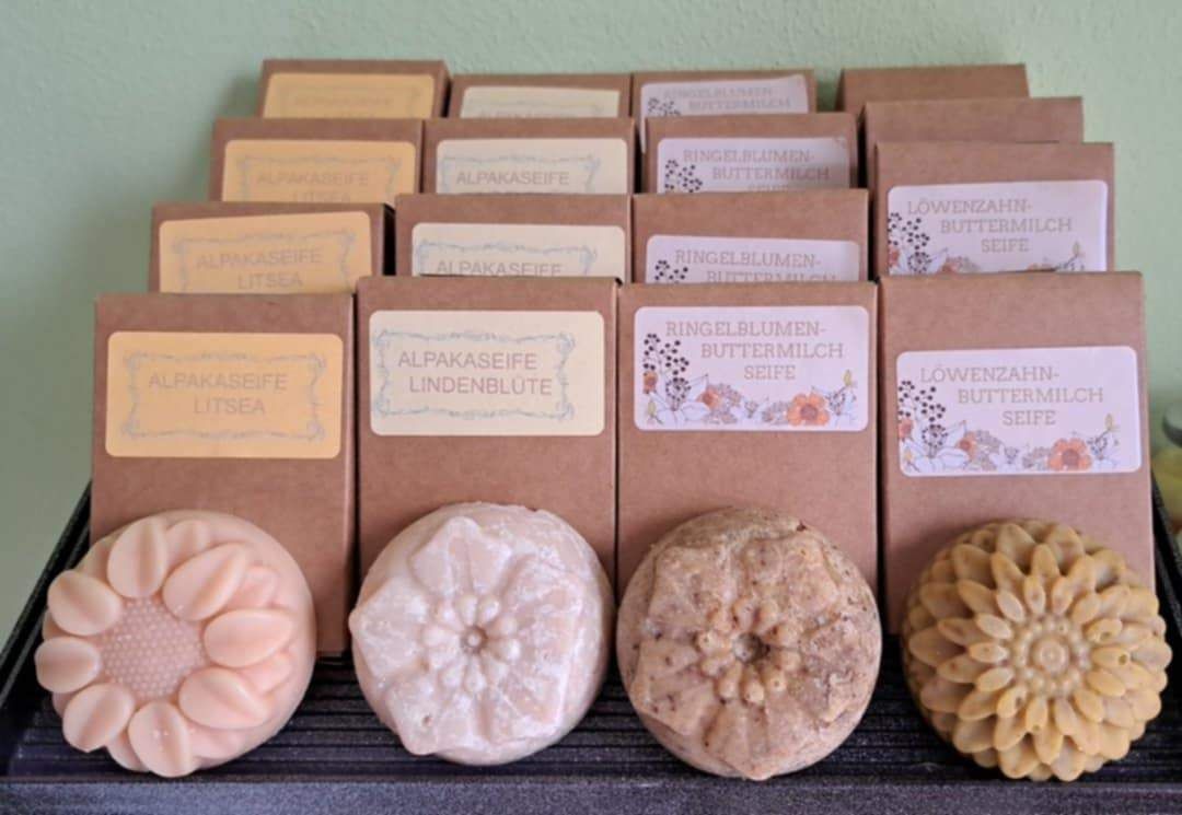 Eine Sammlung von handgemachten Seifen in verschiedenen Formen und Größen, jede in braunes Papier mit gelben Etiketten verpackt. Die Seifen sind mit verschiedenen Zutaten und Typen beschriftet.