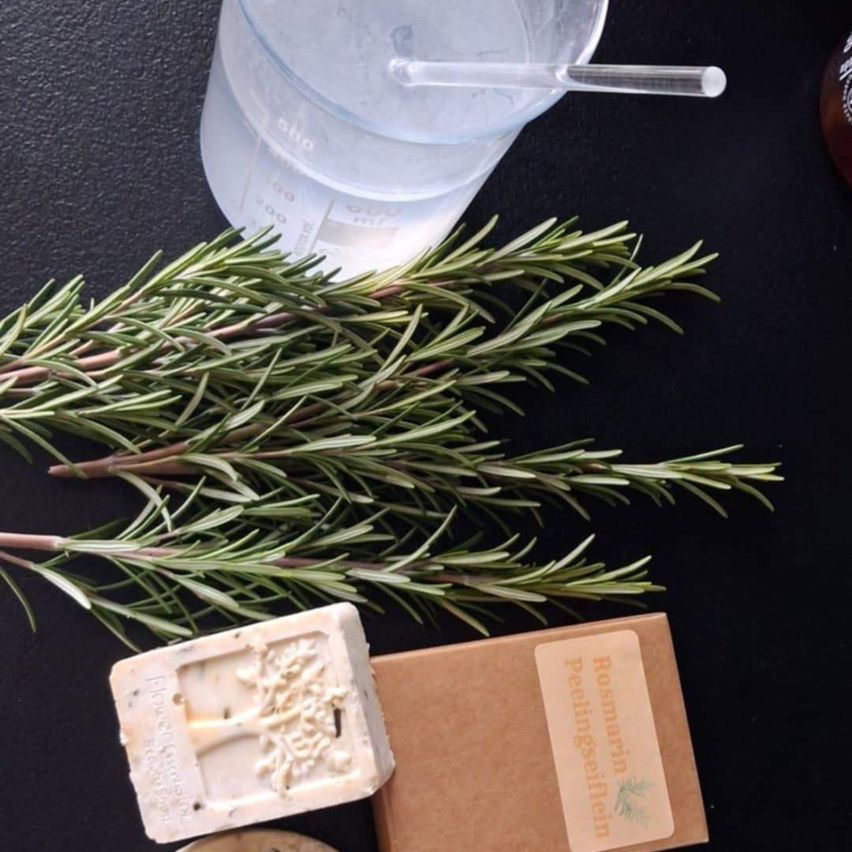 Eine Nahaufnahme einer schwarzen Oberfläche mit einem Glasmessbecher, einem Rosmarinbüschel, einer Seifenbarre und einer kleinen Schachtel mit Etikett.