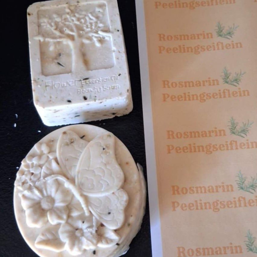 Zwei handgemachte Seifen mit natürlichen Inhaltsstoffen. Eine hat ein Baumdesign, die andere eine Schmetterlingsform. Beide haben die Worte 'Rosmarin Peelingseife' auf der Verpackung.