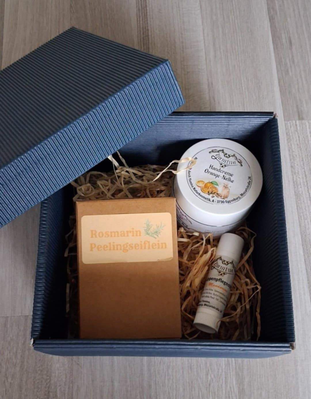 Eine offene blaue Schachtel zeigt verschiedene Produkte im Inneren. Eine kleine Schachtel ist mit Rosmarin Peelingseife beschriftet. Eine Dose orangendufter Creme trägt das Etikett 'Handcreme Orange-Nehle'. Ein kleiner Pflegestift ist ebenfalls enthalten.