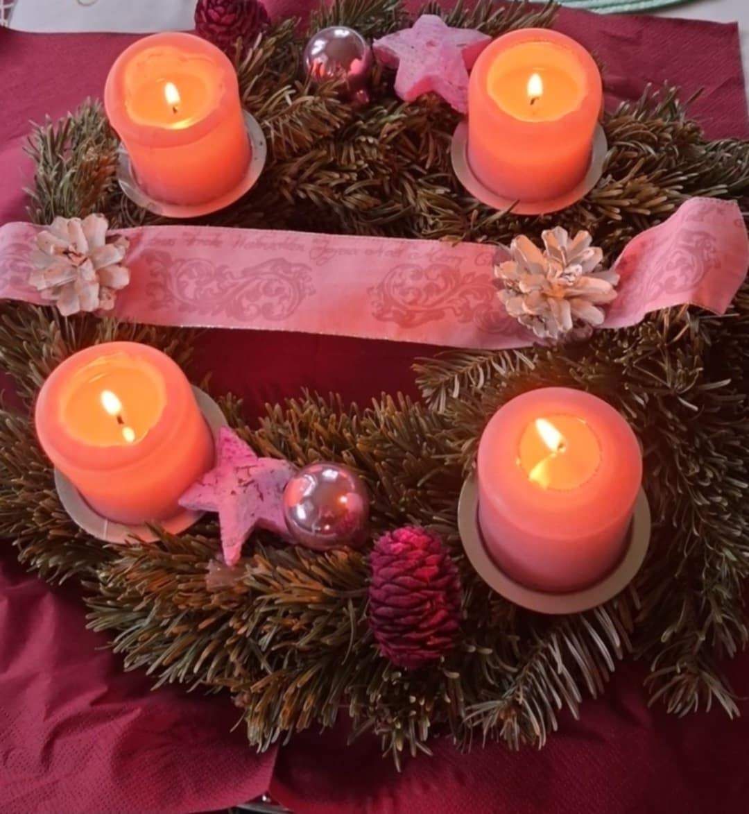 Ein Kranz aus immergrünen Pflanzen hält fünf rosa Kerzen, umgeben von dekorativen Ornamenten und Blumen. Ein rosa Band ist über den Kranz gebunden.