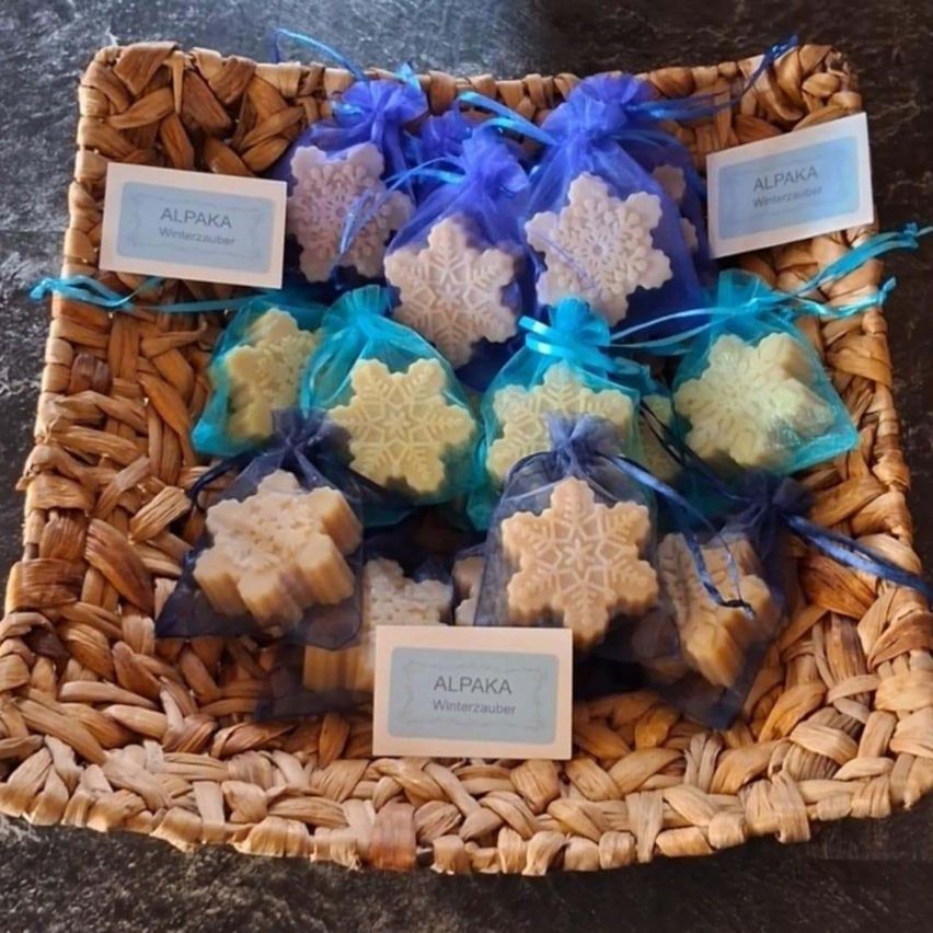 Ein Korb mit blauen Tüten enthält Seifen in Form von Schneeflocken. Jede Tüte hat ein weißes Etikett mit dem Text 'ALPAKA Winterzauber'. Der Hintergrund ist eine dunkle Marmoroberfläche.