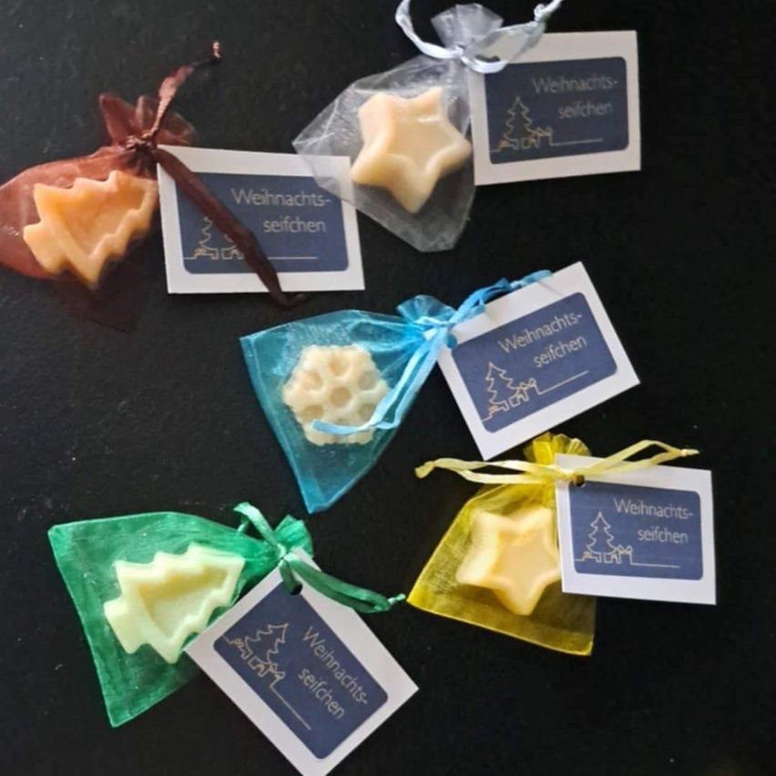 Verschiedene selbstgemachte Weihnachtsseifen in Form von Sternen, Schneeflocken und Bäumen, jede in einem kleinen Plastikbeutel mit Etikett verpackt.
