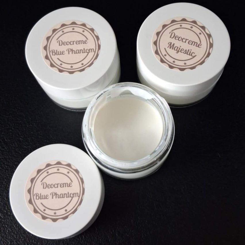 Drei Gläser von 'Decreeme Blue Phantom' auf einer schwarzen Oberfläche. Die Gläser sind weiß mit braunen Etiketten, die den Markennamen tragen. Ein Glas ist geöffnet und zeigt weißen Creme.