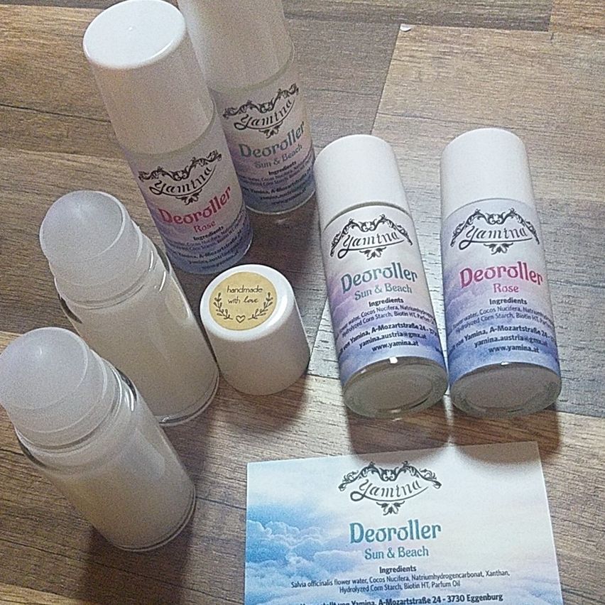 Mehrere Deo-Flaschen liegen auf einer Holzoberfläche. Sie sind mit 'Deoroller' und 'Sonne & Strand' beschriftet. Eine Flasche hat einen Rosenduft. Ein kleines Kärtchen mit den Worten 'Deoroller Sonne & Strand' ist ebenfalls vorhanden.