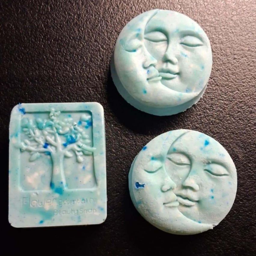 Three soap bars with unique designs on a black surface. The left one has a tree, the middle one has a moon face, and the right one has a crescent moon face.