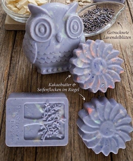 A wooden table displays various handmade soaps. An owl-shaped soap with lavender and cocoa butter, a flower-shaped soap with lavender petals, and a tree-shaped soap with lavender essential oil.