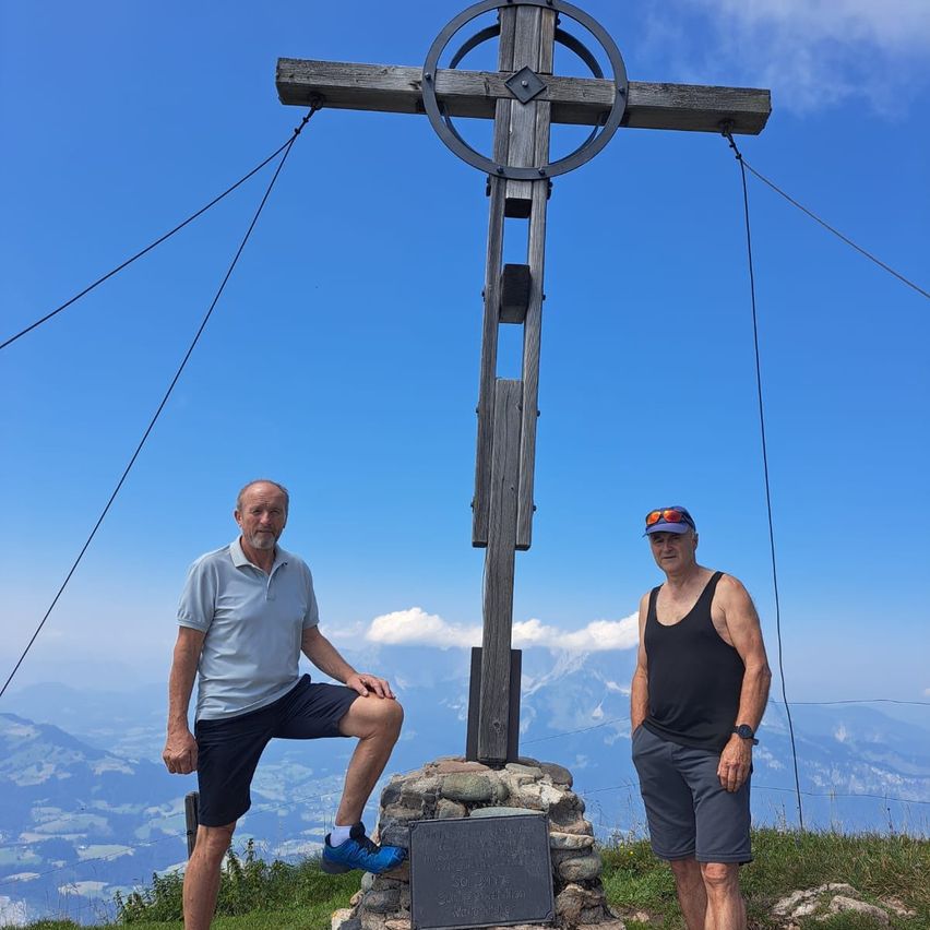 Zwei Männer stehen auf einem Berggipfel, einer trägt ein blaues Hemd und der andere ein schwarzes Tanktop. Sie posieren für ein Foto vor einem Holzkreuz.