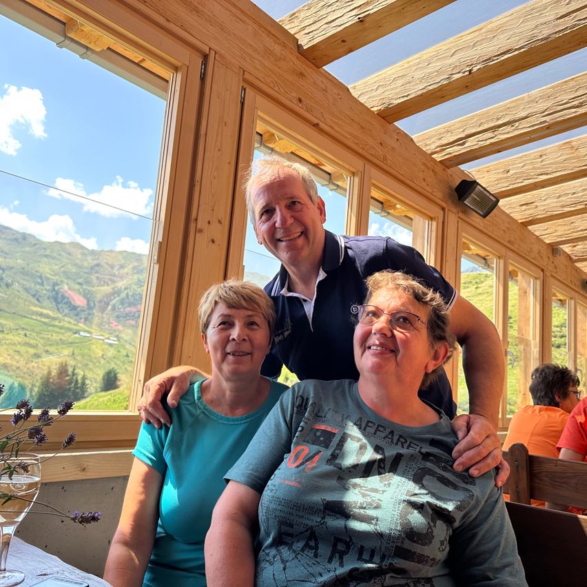 Drei Personen, zwei Frauen und ein Mann, sitzen in einer Hütte mit Holzwänden und großen Fenstern. Die Hütte hat Aussicht auf einen Berg mit Grünflächen. Die Frau links trägt ein blaues Shirt, die Frau rechts ein graues Shirt mit Text. Der Mann steht hinter ihnen und lächelt.