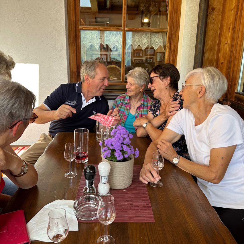 Mehrere Personen sitzen um einen Tisch und lachen. Eine Frau hält ein Weinglas, eine andere eine Blume. Ein Mann gestikuliert und lächelt. Eine Vase mit lila Blumen steht auf dem Tisch.