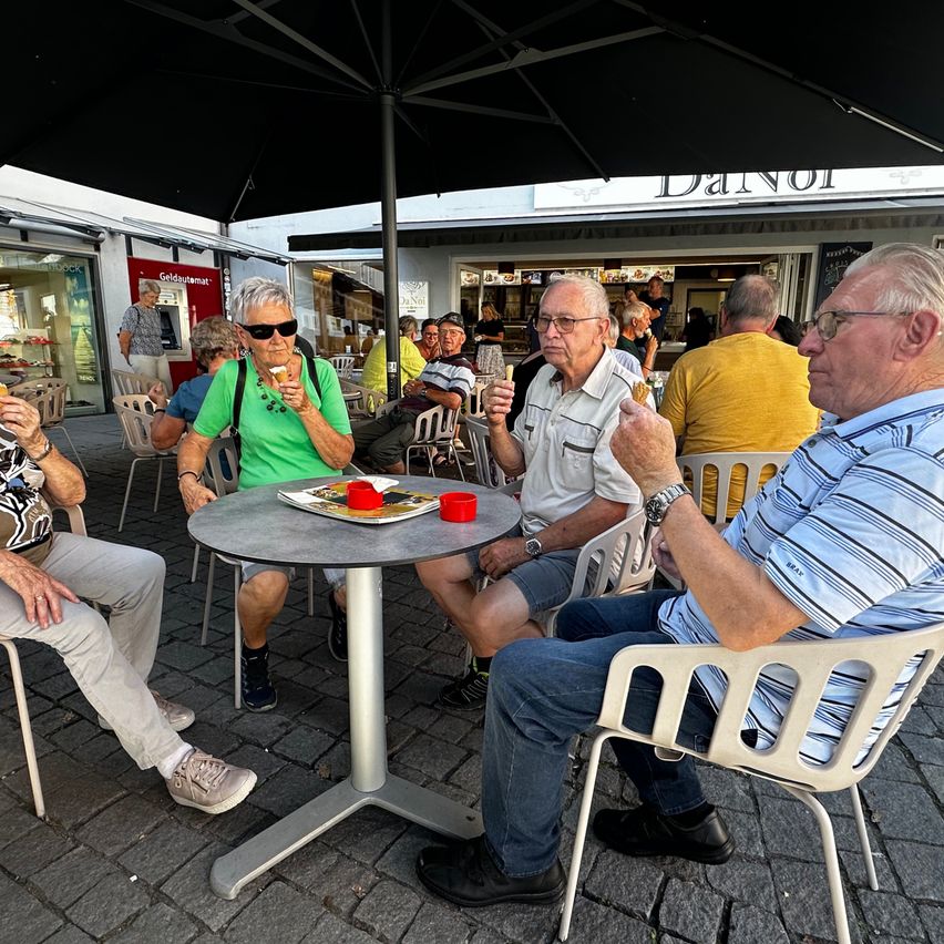 Eine Gruppe älterer Menschen sitzt draußen an einem Tisch, isst Eis und plaudert. Auf dem Tisch steht ein Teller mit einem roten Becher und einer Zeitschrift. Sie sitzen unter einem großen Schirm.
