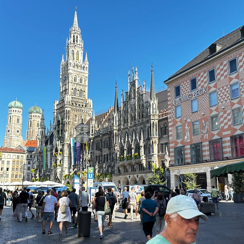 Ein belebter Stadtplatz mit einer großen gotischen Kathedrale im Hintergrund und einem modernen Gebäude mit dem Namen Ludwig Beck. Menschen gehen herum, einige tragen Taschen.