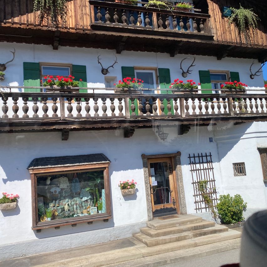 Ein traditionelles europäisches Haus mit einem hölzernen Balkon, der mit roten Blumen und Hirschgeweihen geschmückt ist, einem Schaufenster, das verschiedene Gegenstände präsentiert, und einer Holztür mit einem Metalltor.