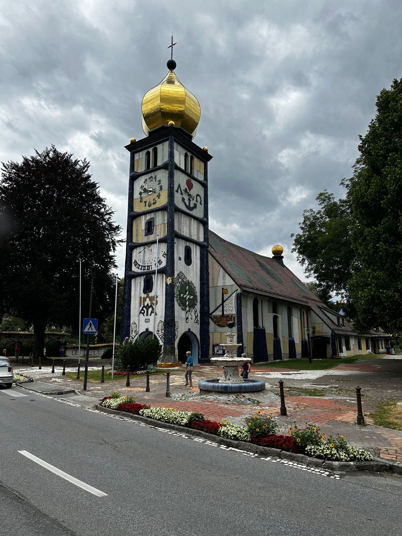 A church with a golden dome and clock tower stands in front of a fountain, with two people walking nearby.