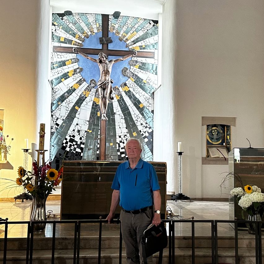 An older man stands in front of a church altar, holding a book. Behind him is a colorful mosaic of Jesus on the cross.