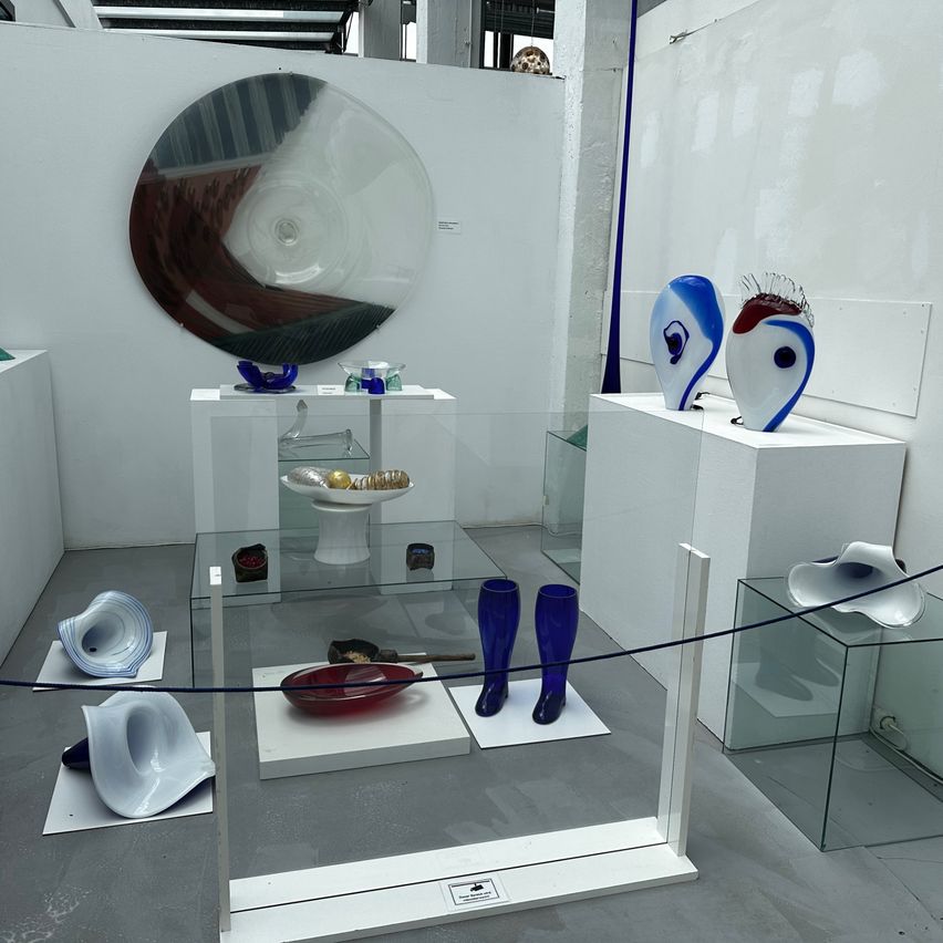 An indoor gallery displays various glass sculptures, including a large circular piece, blue vases, and abstract forms, all on white pedestals and glass tables.