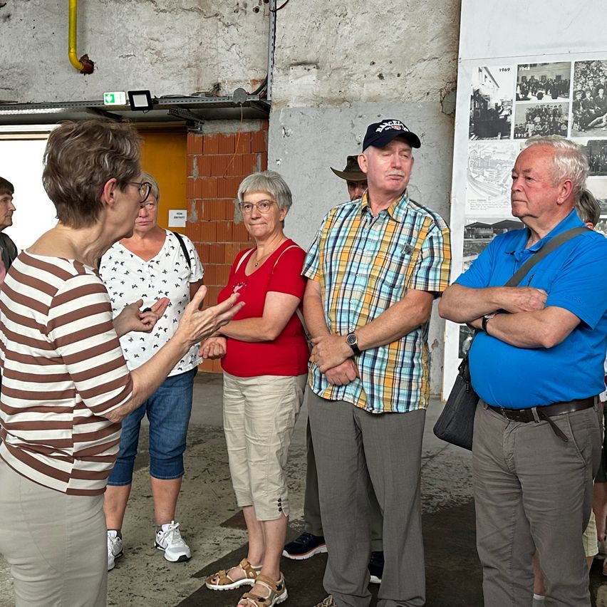 A group of people, including an elderly man and woman, are standing in a room with a brick wall and a photo collage on the wall. They are engaged in a conversation.
