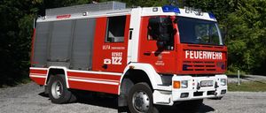 Ein rot-weißer Feuerwehrwagen mit der Bezeichnung ULFA 02682 122 und Notleuchten auf dem Dach, geparkt auf Kies unter Bäumen.