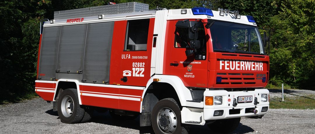 Ein rot-weißer Feuerwehrwagen mit der Bezeichnung ULFA 02682 122 und Notleuchten auf dem Dach, geparkt auf Kies unter Bäumen.