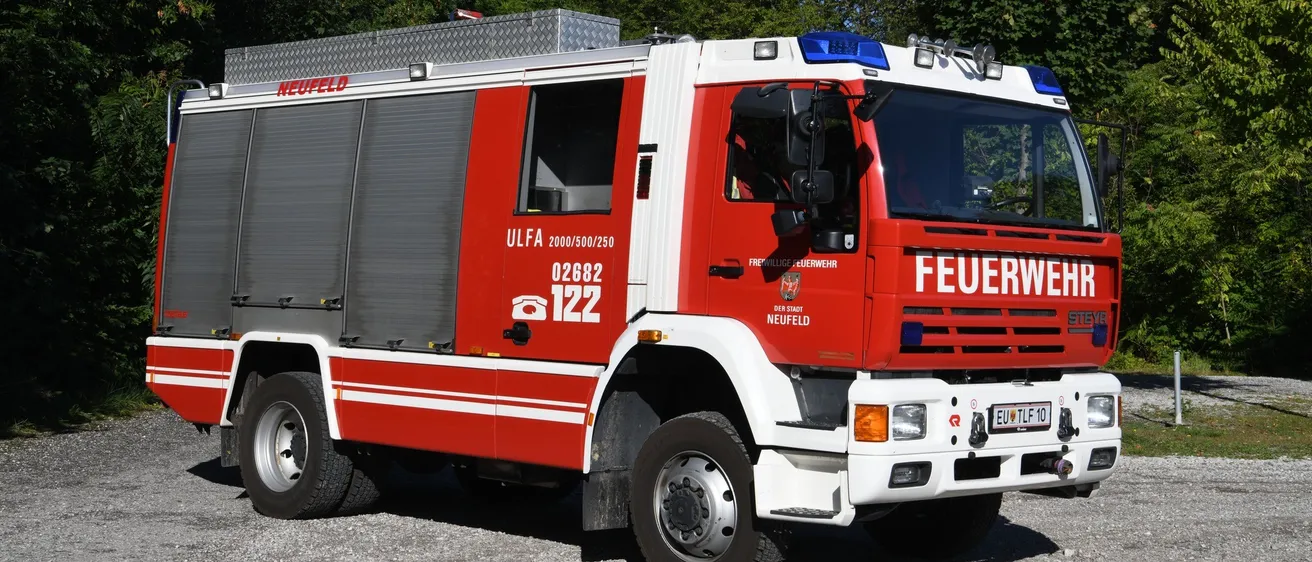 Ein rot-weißer Feuerwehrwagen mit der Bezeichnung ULFA 02682 122 und Notleuchten auf dem Dach, geparkt auf Kies unter Bäumen.