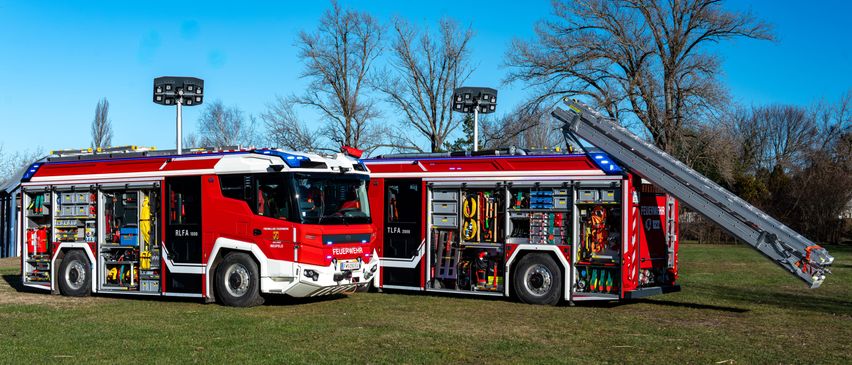 Zwei Feuerwehrfahrzeuge sind auf einem Rasenfeld geparkt, beide mit ausgeklappten Leitern. Die Fahrzeuge sind rot mit weißen Streifen und mit 'FEUERWEHR' beschriftet.