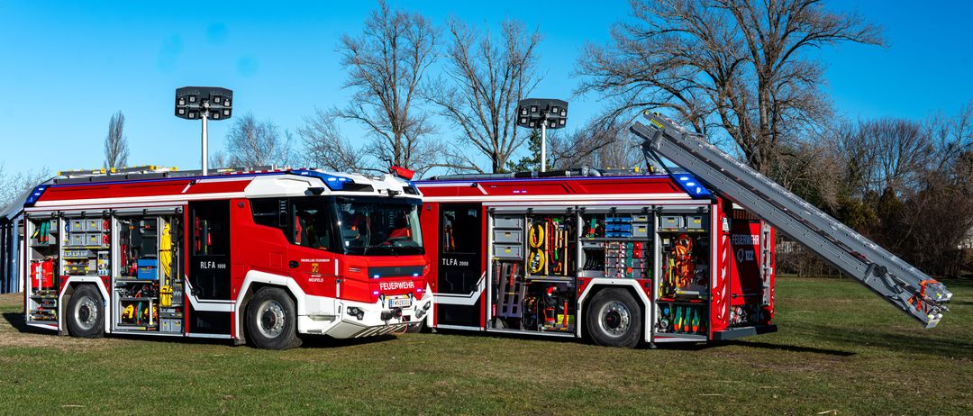 Zwei Feuerwehrfahrzeuge sind auf einem Rasenfeld geparkt, beide mit ausgeklappten Leitern. Die Fahrzeuge sind rot mit weißen Streifen und mit 'FEUERWEHR' beschriftet.