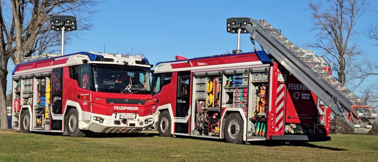 Zwei rote Feuerwehrfahrzeuge sind auf einem Rasenfeld mit offenen Türen geparkt. Ein Fahrzeug hat seine Leiter ausgefahren.