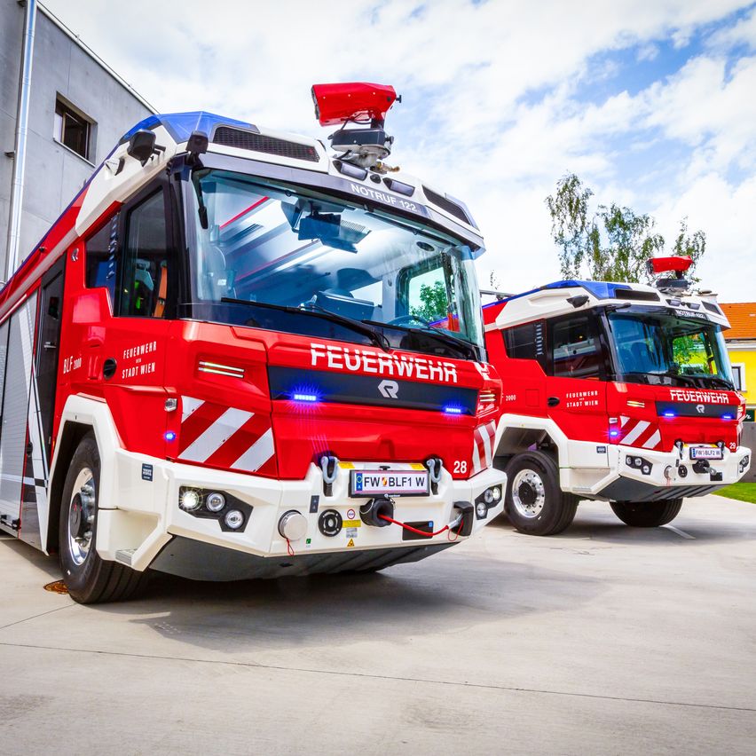 Zwei rote und weiße Feuerwehrfahrzeuge sind in einer Garage geparkt. Die Front der Fahrzeuge trägt die Aufschrift 'FEUERWEHR' und die Nummer '28'. Der Himmel ist blau mit einigen Wolken.
