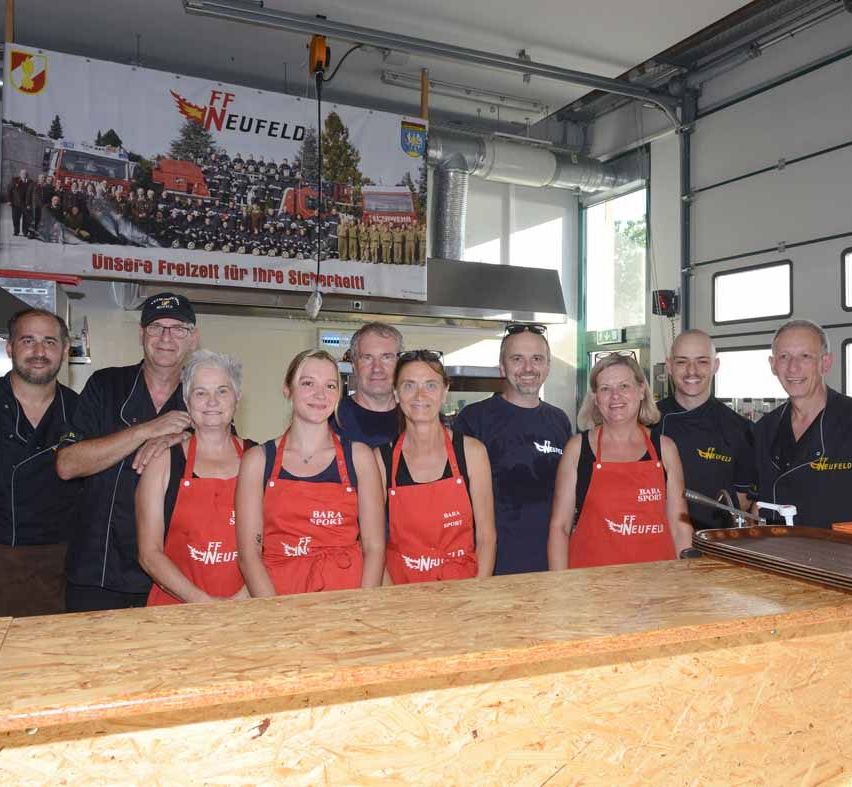 Eine Gruppe von Menschen in roten Schürzen posiert für ein Foto in einer Küche. Über ihnen steht auf dem Banner FF Neufeld mit Bildern von Feuerwehrleuten.