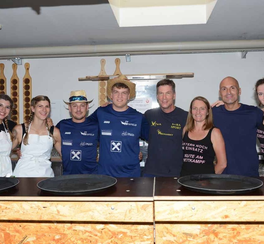 Eine Gruppe von Menschen steht hinter einer Theke in einem Raum mit Holzwänden und -decke und posiert für ein Foto. Sie tragen verschiedene T-Shirts mit Logos und eine Frau hat eine weiße Schürze.