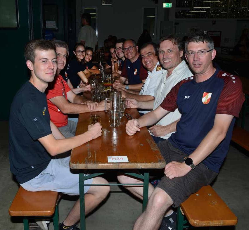 Eine Gruppe von Menschen sitzt um einen Holztisch in einem Restaurant. Sie haben Gläser vor sich. Sie lächeln und scheinen für ein Foto zu posieren. Der Tisch hat eine Nummer darauf. Das Restaurant hat ein gedämpftes Licht.