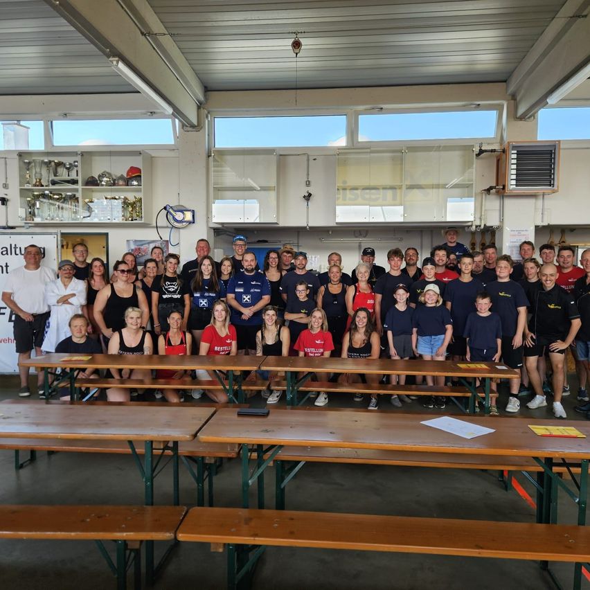 Ein Gruppenfoto von Personen in einem Raum mit Tischen, Bänken und einer Decke mit Lichtern. Einige tragen Hüte und Brillen, und Trophäen sind an Regalen. Papiere liegen auf den Tischen.