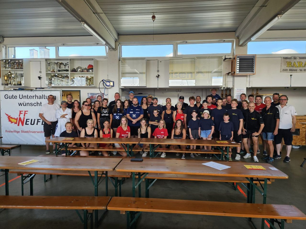 Ein Gruppenfoto von Personen in einem Raum mit Tischen, Bänken und einer Decke mit Lichtern. Einige tragen Hüte und Brillen, und Trophäen sind an Regalen. Papiere liegen auf den Tischen.
