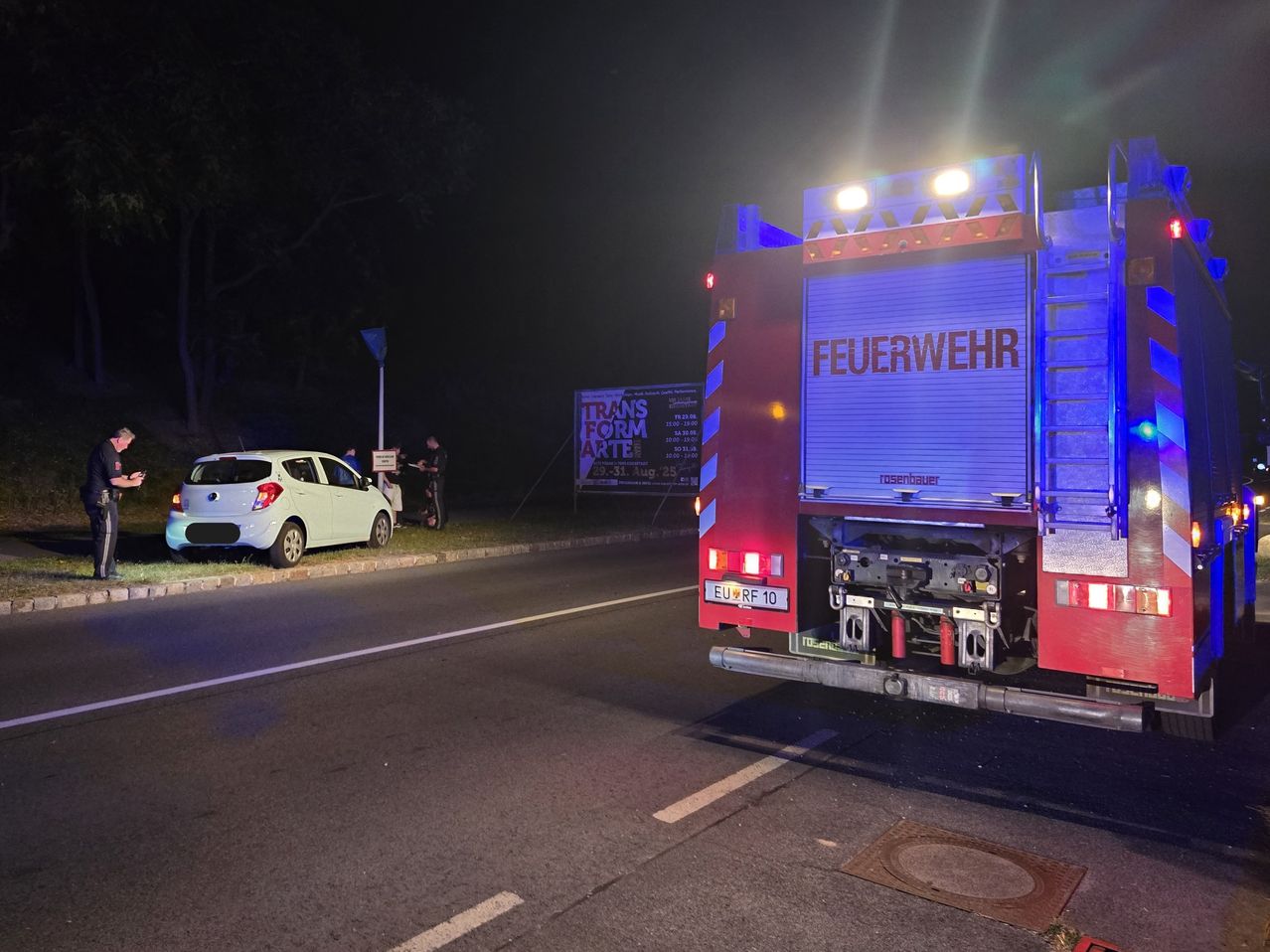 Ein weißer Wagen ist nachts am Straßenrand geparkt. Dahinter steht ein Feuerwehrwagen, und zwei Personen stehen in der Nähe eines Schildes. Das Gebiet ist dunkel, mit Bäumen auf der linken Seite und Straßenlaternen, die die Szene beleuchten.