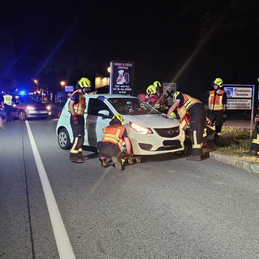 Rettungskräfte kümmern sich bei Nacht um ein beschädigtes Auto auf der Straße. Sie tragen reflektierende Westen und Helme, und einige schieben das Auto von der Straße weg.