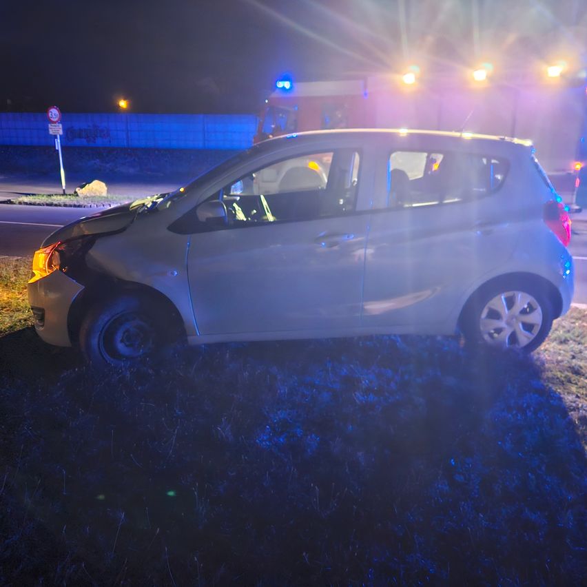 Ein weißer Wagen mit vorderseitiger Beschädigung steht bei Nacht am Straßenrand, im Hintergrund sind Notfallleuchten zu sehen.
