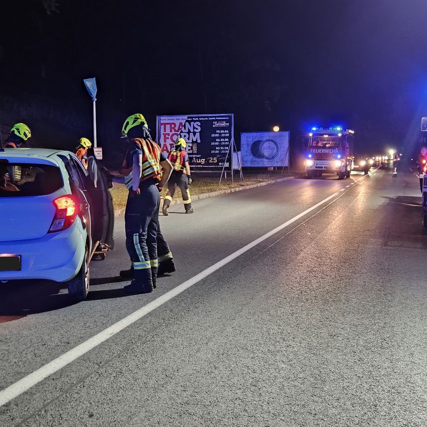 Feuerwehrleute in Schutzkleidung helfen einem Auto auf einer Straße bei Nacht. Ein Feuerwehrwagen mit blinkenden Lichtern ist in der Nähe geparkt.
