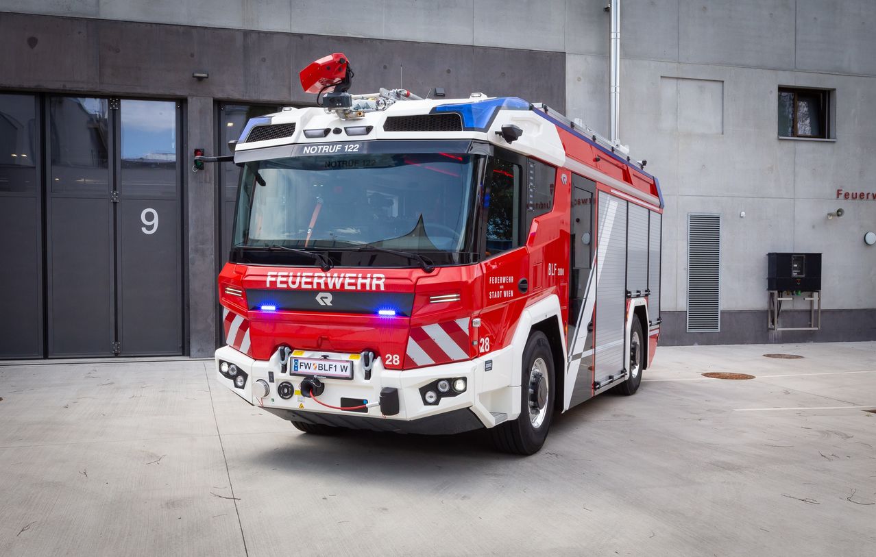 Ein roter und weißer Feuerwehrwagen ist vor einem Gebäude geparkt. Der Wagen hat ein Nummernschild 'FW BLFI W 28'. Er ist mit 'Feuerwehr' beschriftet und hat Notfallleuchten.