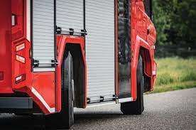Ein roter Feuerwehrwagen steht am Straßenrand, mit geöffneter Hecktür. Der Körper des Wagens besteht aus Metall, und hinten ist ein Ersatzreifen montiert.