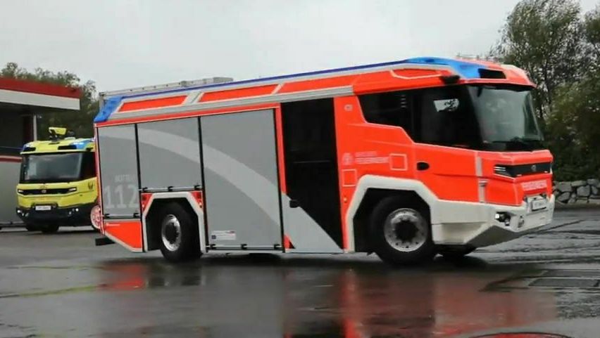 Ein roter und weißer Feuerwehrwagen mit der Nummer 112 ist in einem nassen Bereich geparkt, möglicherweise an einer Tankstelle. Ein weiteres gelbes und blaues Notfallfahrzeug ist dahinter geparkt.