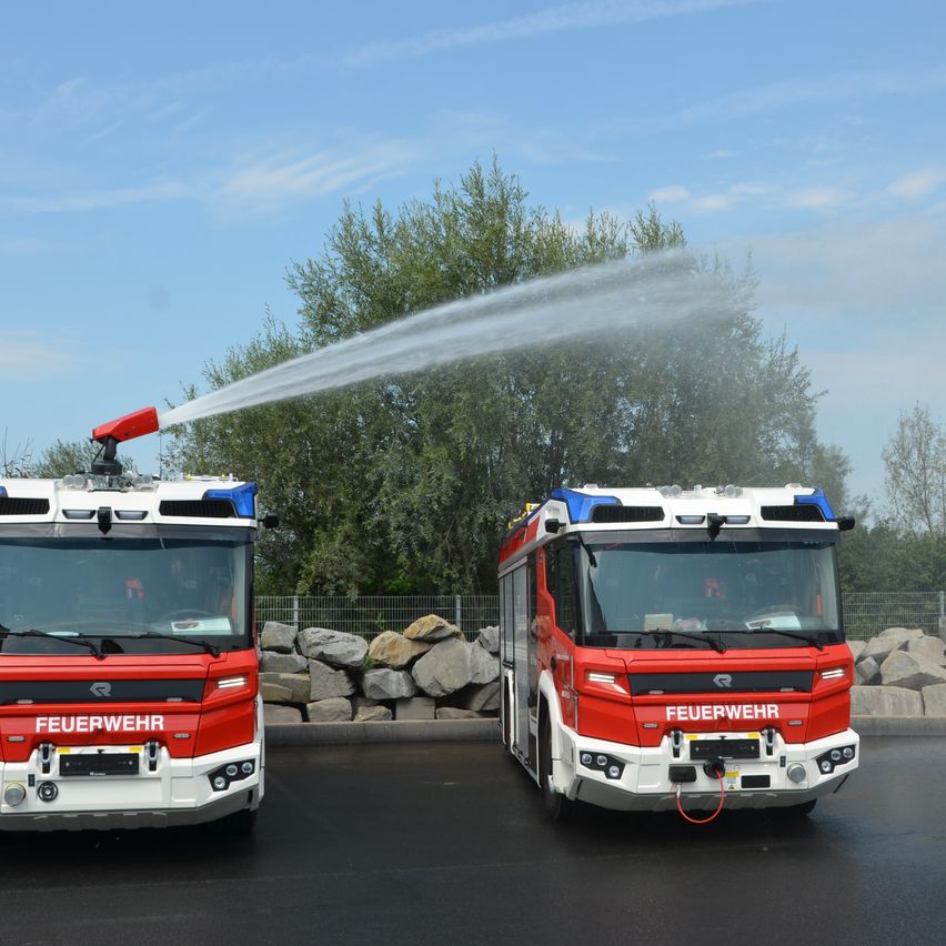 Zwei Feuerwehrfahrzeuge sprühen Wasser aus ihren Schläuchen. Sie stehen auf einer Straße mit einer Steinmauer und Bäumen dahinter. Der Himmel ist blau mit einigen Wolken.