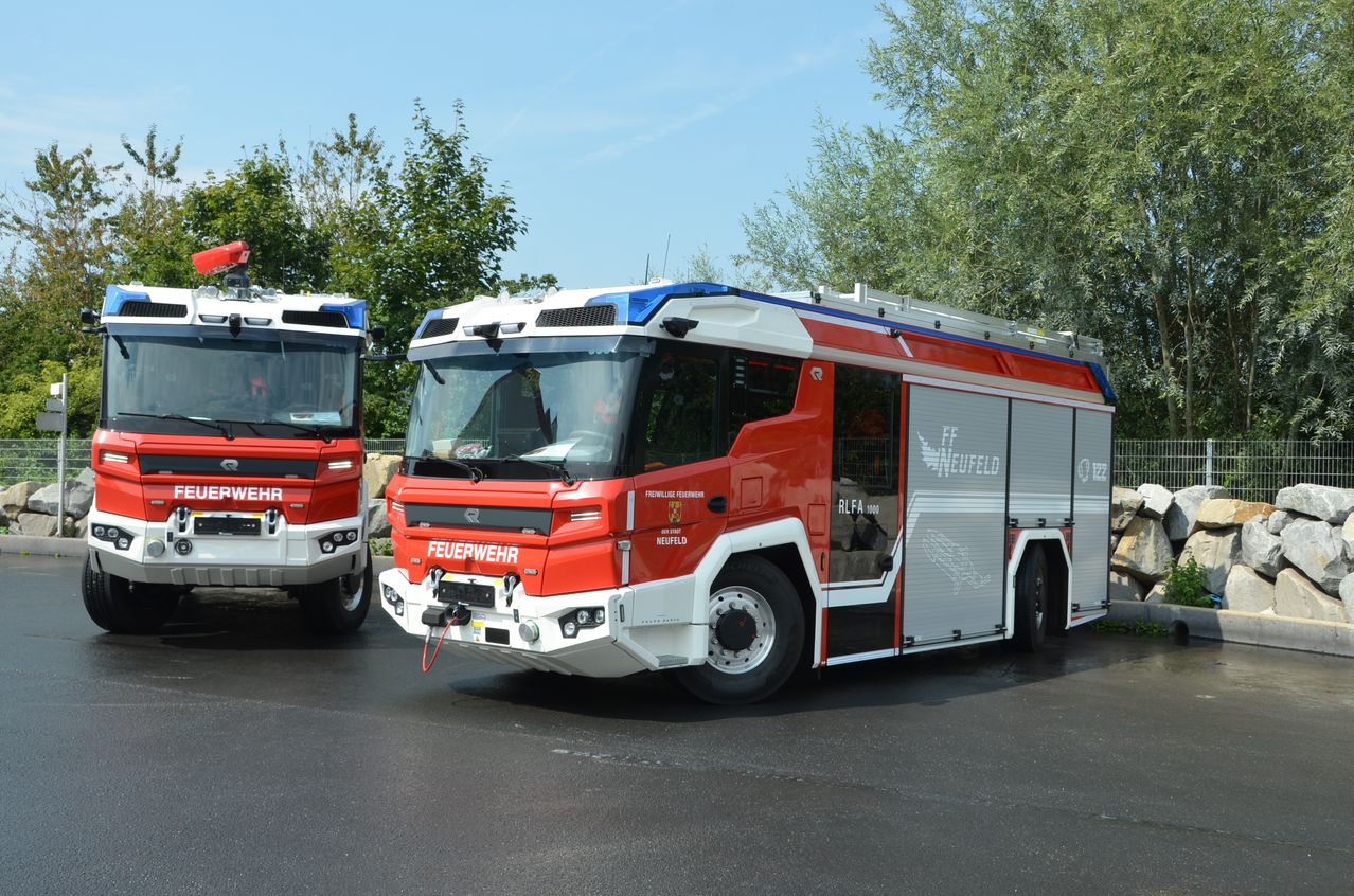Zwei rote und weiße Feuerwehrfahrzeuge stehen in einem Außenbereich. Eines hat ein 'Feuerwehr'-Schild und das andere ein 'FF'. Bäume und eine Steinmauer im Hintergrund.