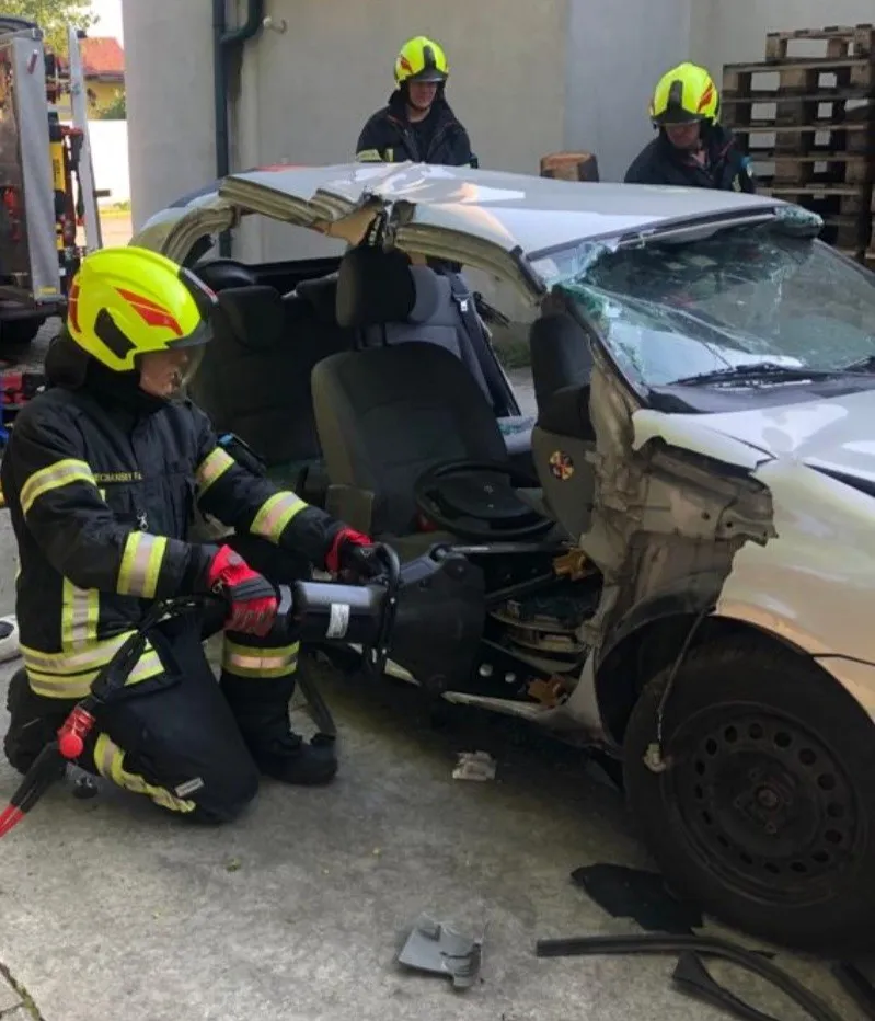 Ein Feuerwehrmann benutzt ein Werkzeug, um in ein beschädigtes Fahrzeug zu schneiden. Zwei andere Feuerwehrleute stehen hinter dem Fahrzeug, einer von ihnen hält einen Feuerlöscher.