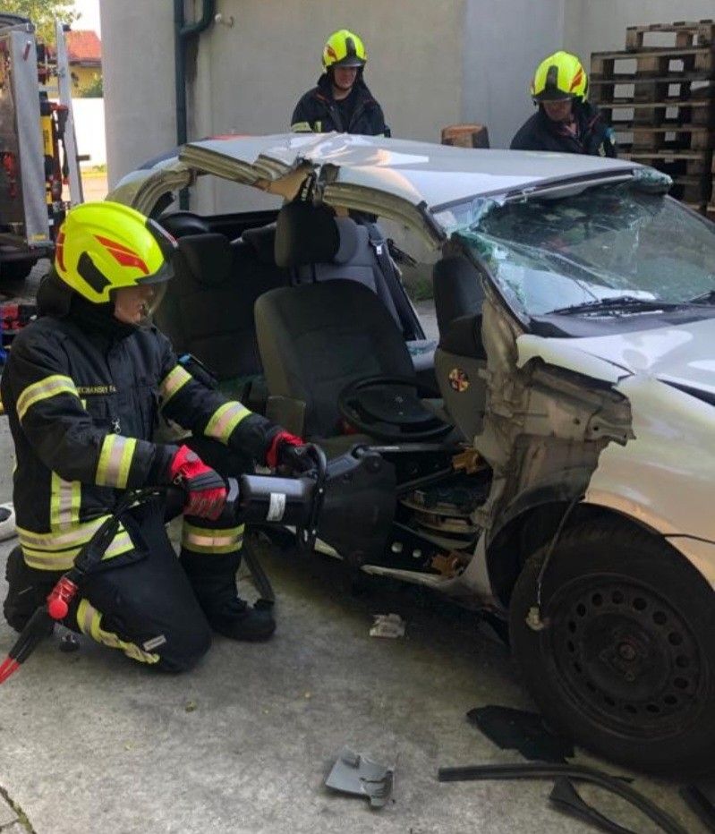 Ein Feuerwehrmann benutzt ein Werkzeug, um in ein beschädigtes Fahrzeug zu schneiden. Zwei andere Feuerwehrleute stehen hinter dem Fahrzeug, einer von ihnen hält einen Feuerlöscher.