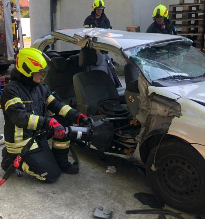 Ein Feuerwehrmann benutzt ein Werkzeug, um in ein beschädigtes Fahrzeug zu schneiden. Zwei andere Feuerwehrleute stehen hinter dem Fahrzeug, einer von ihnen hält einen Feuerlöscher.