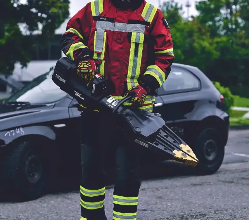 Ein Feuerwehrmann in einem roten Uniform mit gelben und grauen Streifen steht neben einem schwarzen Auto und hält ein großes schwarzes und goldenes Werkzeug in den Händen.