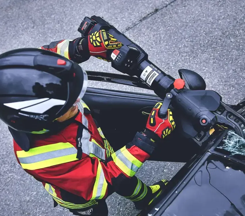 Ein Feuerwehrmann benutzt ein Werkzeug, um die Tür eines Autos zu öffnen. Die Person trägt einen Helm, Handschuhe und eine Sicherheitsjacke. Das Auto ist auf der Straße geparkt.
