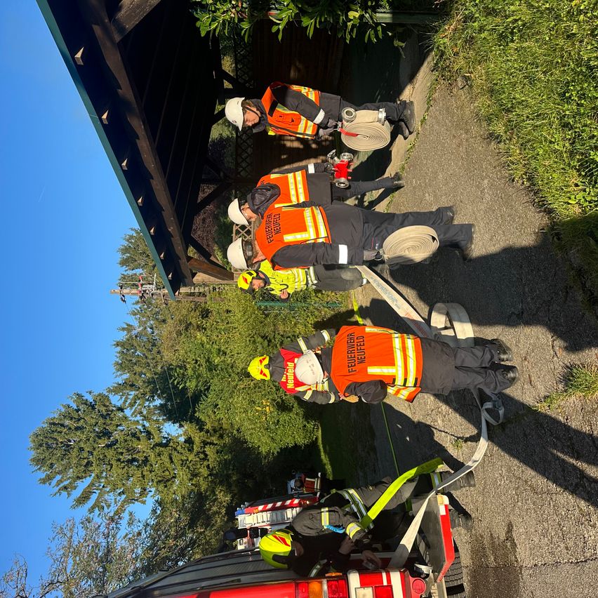 Mehrere Feuerwehrleute sind auf einer Straße und bereiten einen Schlauch vor. Sie sind in reflektierenden Westen und Helmen gekleidet. In der Nähe ist ein Feuerwehrwagen geparkt. Bäume und Grünfläche umgeben die Szene.