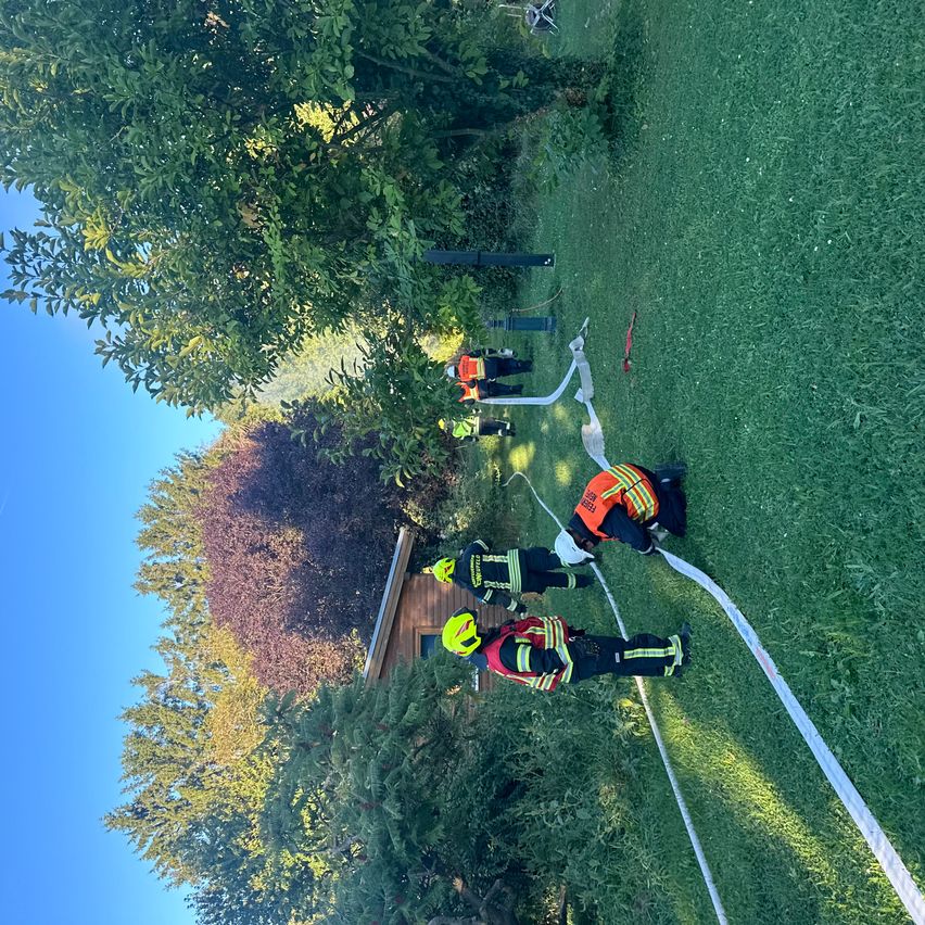 Mehrere Feuerwehrleute in Warnschutzkleidung arbeiten auf einem Rasen. Sie verlegen eine lange Schlange. Dahinter steht ein Haus mit einem schrägen Dach. Bäume umgeben das Gebiet.